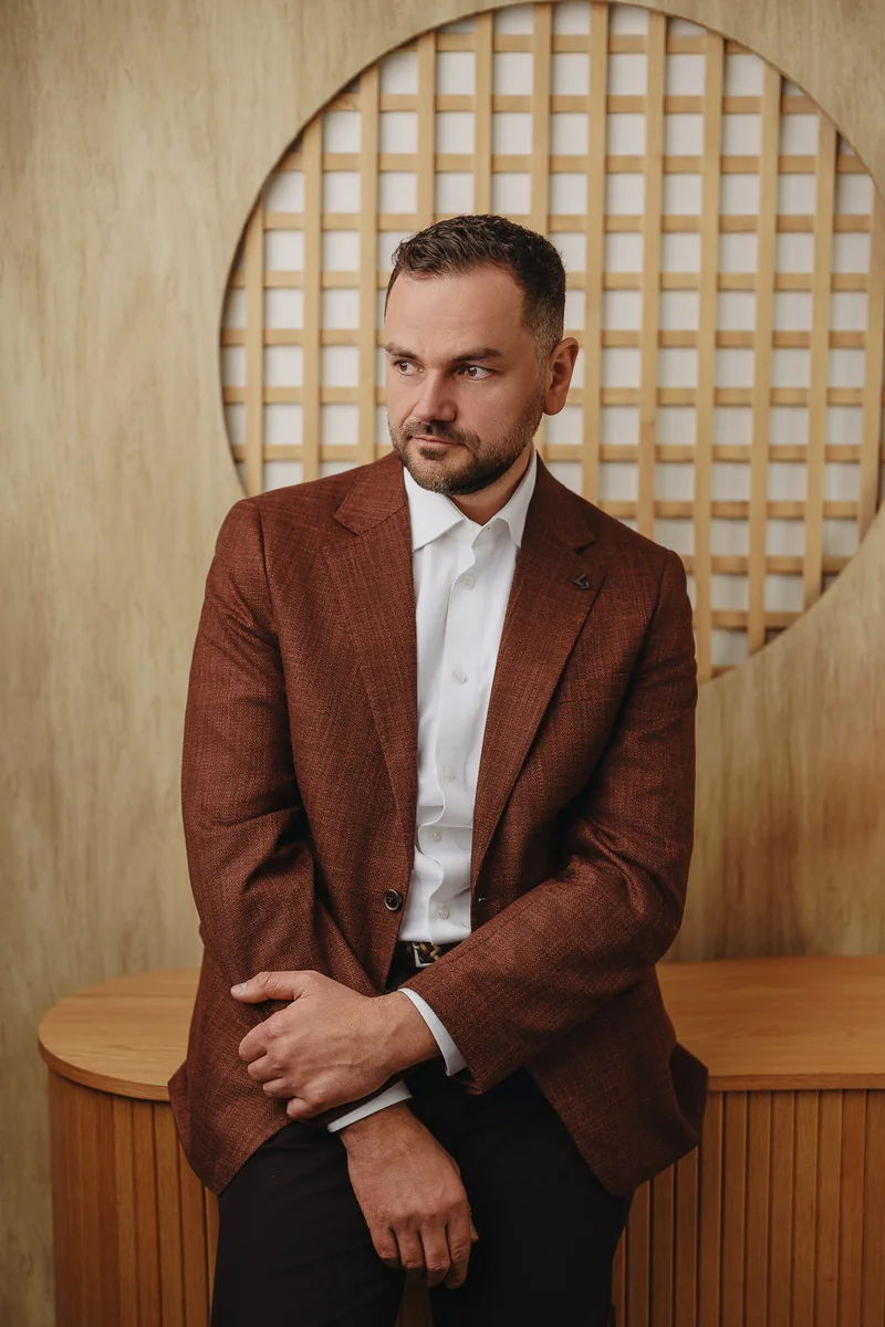 Marek Wolan w brązowej marynarce, siedzący w jasnym wnętrzu